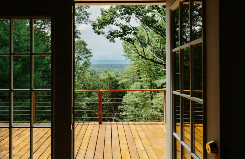 View from an open door showcasing a wooden deck surrounded by lush green trees and distant mountains.