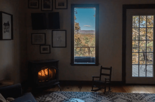 Cozy living room with a wood stove, framed art, and a view of trees and mountains through a window and glass door.