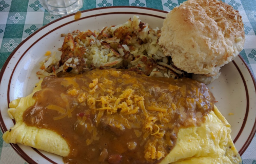 A plate featuring a cheese omelet topped with chili, hash browns, and a biscuit on a checkered tablecloth.