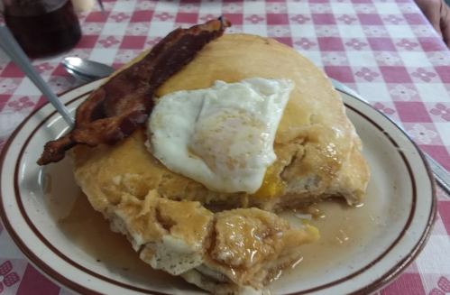 A large pancake topped with a fried egg and a slice of bacon, served on a plate with syrup.