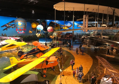 A vibrant museum display featuring various vintage aircraft and hot air balloons, with visitors exploring the exhibits.
