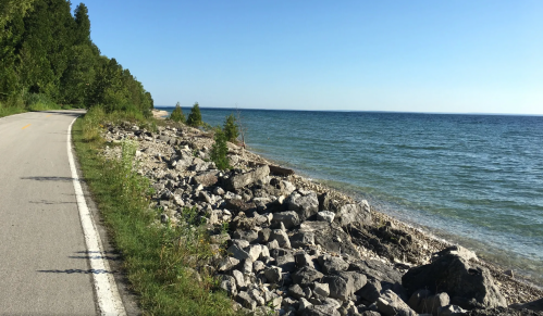 A scenic view of a winding road alongside a rocky shoreline and calm blue water under a clear sky.