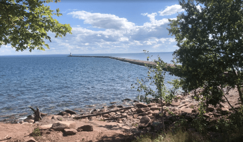A serene view of a pier extending into a calm lake, surrounded by trees and rocky shoreline under a blue sky.
