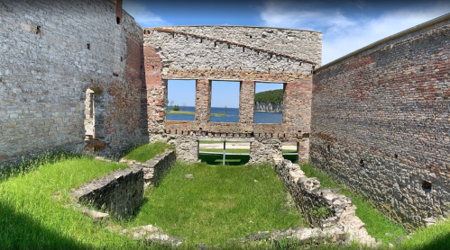 Ruins of a stone building with grassy interior, overlooking a body of water and trees in the background.