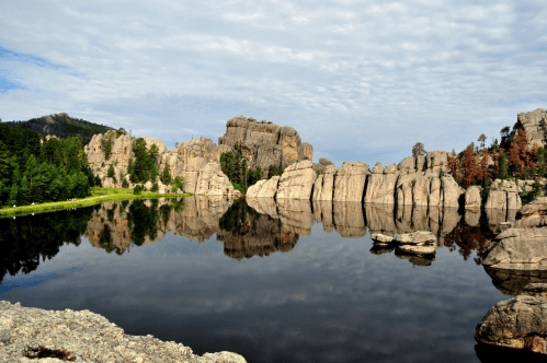 A serene lake reflects rocky cliffs and trees under a cloudy sky, creating a tranquil natural landscape.