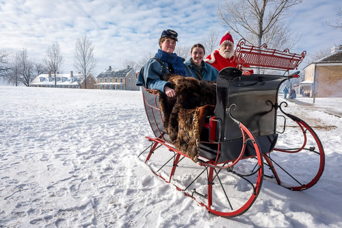 A man in a Santa suit sits in a sleigh with two others, surrounded by snow and historic buildings in the background.