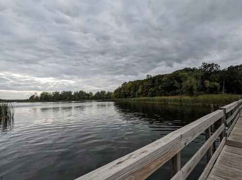 A calm lake under a cloudy sky, with a wooden dock extending into the water and lush greenery along the shore.