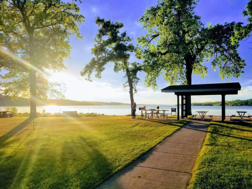 A serene lakeside park at sunset, featuring trees, picnic tables, and a pathway leading to the water.