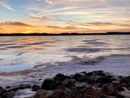 A serene winter landscape featuring a frozen lake at sunset, with colorful clouds reflecting on the icy surface.