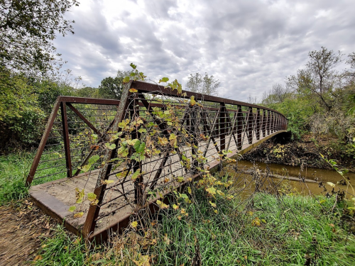 A metal bridge spans a small creek, surrounded by greenery and overcast skies.