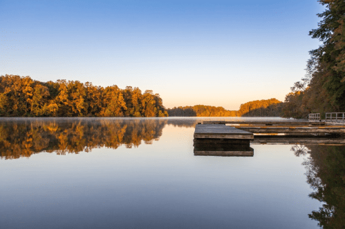 Serene lake at sunrise, with wooden docks and colorful trees reflecting on calm water.