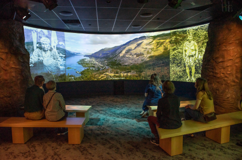 Visitors sit on benches in a circular room, watching historical images projected on large screens.
