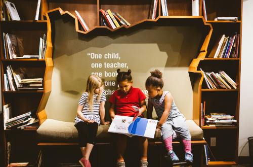 Three children sit on a bench in a library, reading a book together, surrounded by shelves of books.
