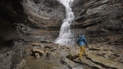 A person in a blue jacket and yellow pants walks on rocks near a waterfall in a rocky, forested area.