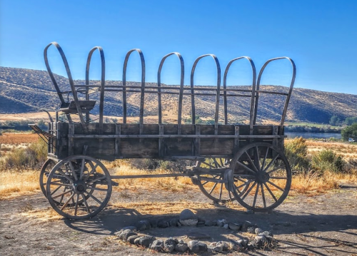 A vintage wooden wagon with metal arches, set against a scenic landscape of hills and a clear blue sky.