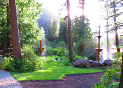 A serene forest scene with two fountains surrounded by lush greenery and tall trees, illuminated by soft sunlight.