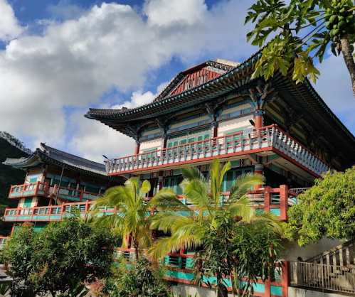 A colorful traditional building with ornate architecture, surrounded by lush greenery and a blue sky with clouds.