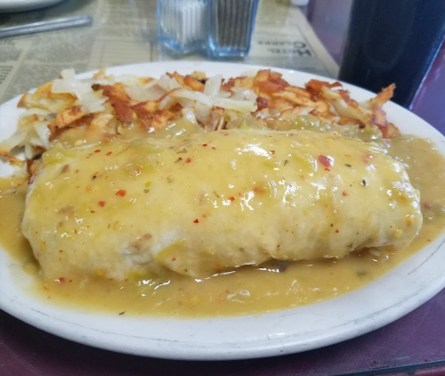 A plate of a large burrito smothered in green sauce, served with crispy hash browns on the side.