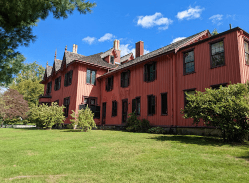 A large, red Victorian-style house with multiple windows, surrounded by green grass and trees under a blue sky.