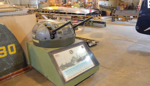 A military display featuring a turret with cannons, set in an aircraft hangar with various aircraft parts in the background.