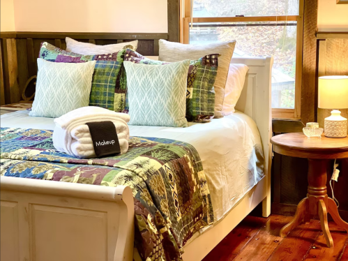 Cozy bedroom with a white bed, colorful quilt, decorative pillows, and a bedside table with a lamp.