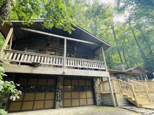 A rustic wooden cabin with a balcony, surrounded by trees, featuring a stone garage and a wooden deck.