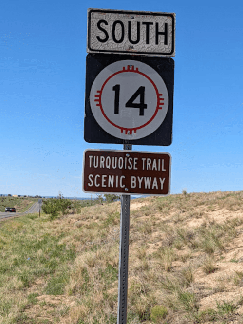Sign indicating South on Route 14, with a brown sign for the Turquoise Trail Scenic Byway. Clear blue sky in the background.
