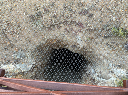 A dark tunnel entrance covered by a metal mesh, with rocky surroundings and rusted metal beams in the foreground.