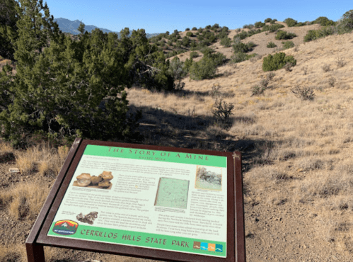 A sign titled "The Story of a Mine" in Cerrillos Hills State Park, with a dry landscape and mountains in the background.