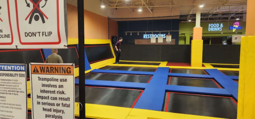 A trampoline park with safety signs, featuring colorful trampolines and a person jumping in the background.