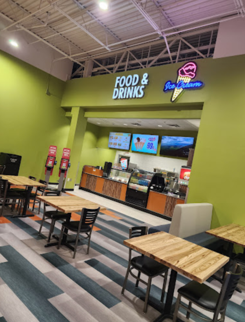 A brightly lit food and drinks counter with tables and chairs in a modern setting, featuring a colorful ice cream sign.