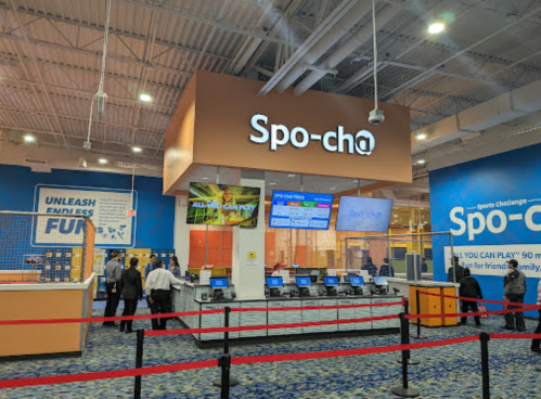 Interior of a sports facility with a reception area, digital screens, and a red queue barrier. "Spo-cho" sign visible.