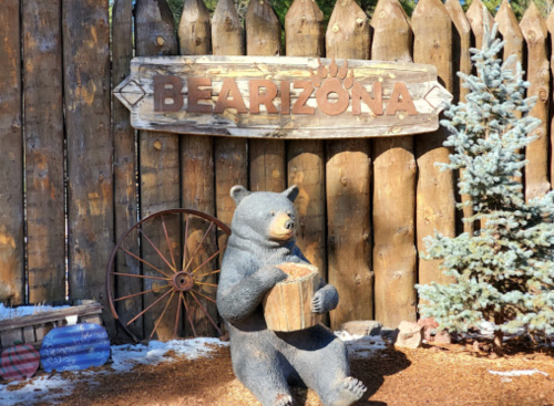 A bear statue holding a log sits in front of a wooden sign that reads "BEARIZONA," surrounded by a rustic fence and trees.