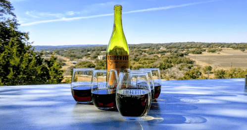 A bottle of wine surrounded by glasses on a table, with a scenic view of rolling hills in the background.