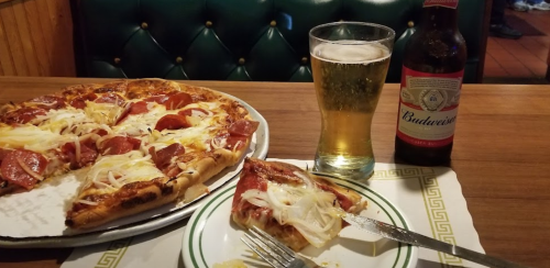 A slice of pepperoni pizza with onions, a glass of beer, and a bottle of Budweiser on a wooden table.