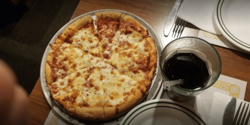 A freshly baked pizza with cheese and pepperoni, sliced into six pieces, next to a glass of dark soda on a table.