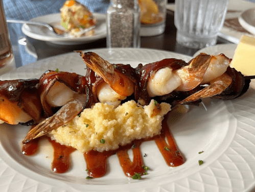 Grilled shrimp skewers wrapped in bacon, served over creamy grits with a drizzle of sauce, on a white plate.