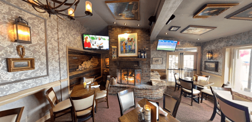 Cozy restaurant interior with a fireplace, artwork on the walls, and TVs showing sports, featuring tables and chairs.