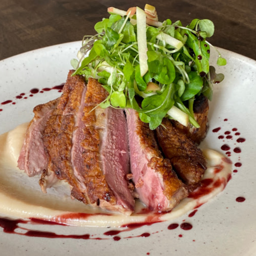 A plate of sliced duck breast topped with greens and apple, served on a creamy base with a berry sauce drizzle.