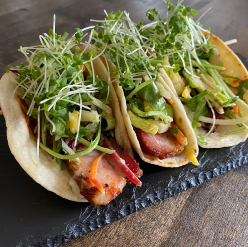 Three tacos filled with grilled meat, fresh vegetables, and sprouts, served on a black slate plate.