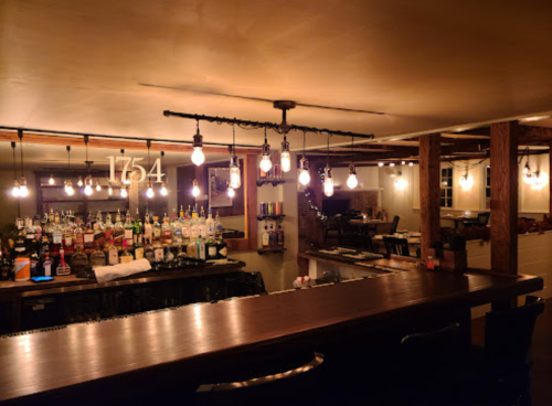 A cozy bar interior with a wooden counter, shelves of liquor, and warm pendant lighting.