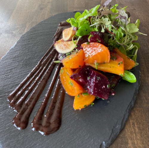 Colorful roasted vegetables, including carrots and beets, garnished with greens and drizzled with a dark sauce on a slate plate.