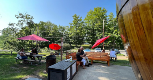 Outdoor seating area with red umbrellas, people relaxing at picnic tables, and a wooden structure in a green park.