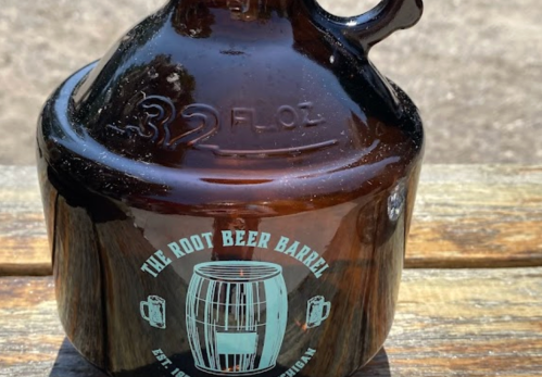 A brown glass jug labeled "The Root Beer Barrel" with a barrel graphic, sitting on a wooden surface.