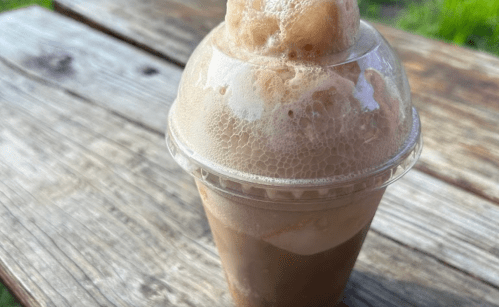 A frothy root beer float in a clear cup, sitting on a rustic wooden table.