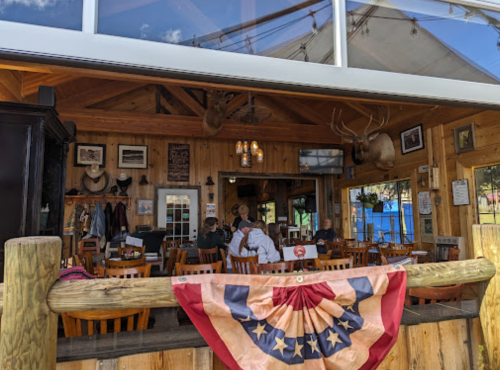 Cozy restaurant interior with wooden decor, antlers on the wall, and patrons dining under a large window.
