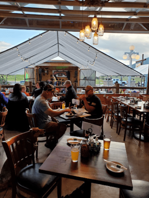 A cozy restaurant scene with diners at wooden tables, enjoying drinks and food under a tented outdoor area.