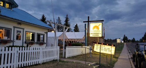 A rustic restaurant and saloon with a blue roof, white picket fence, and a sign, set against a cloudy sky.