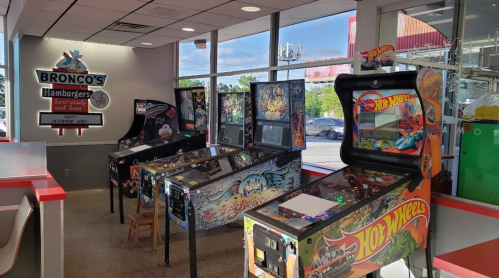 A bright restaurant interior featuring several colorful pinball machines and a sign for Bronco's Hamburgers.