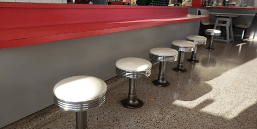 A row of retro diner stools with white seats and metal bases, lined up at a counter with a red and gray design.